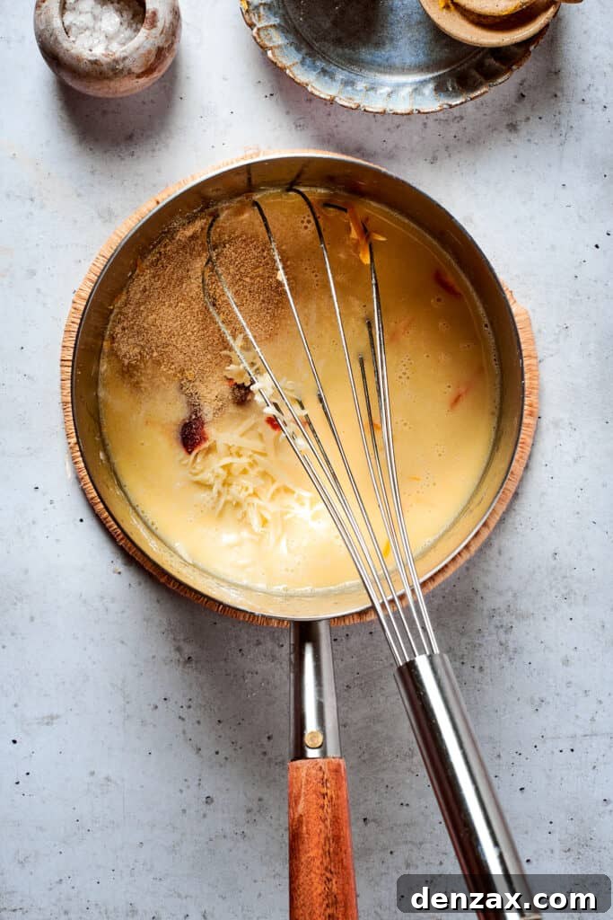 A whisk whisks a cheese sauce in a metal pot.