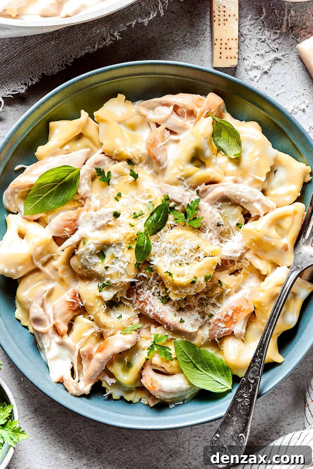 Creamy Chicken Tortellini Alfredo is ready in less than 30 minutes! Rotisserie chicken and tortellini in a cheesy sauce make an easy meal. Close-up overhead shot of chicken tortellini alfredo served in a blue bowl and garnished with basil leaves.