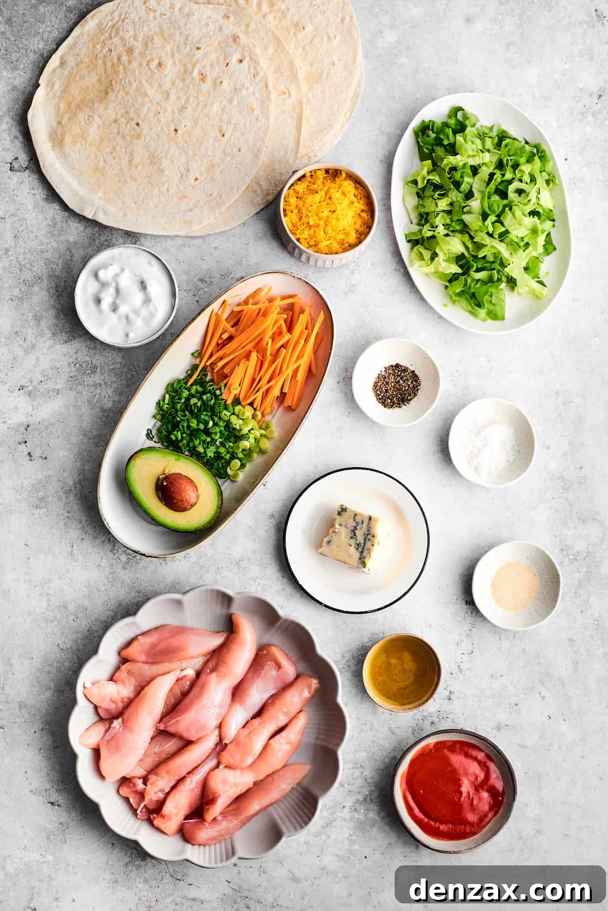 The ingredients for buffalo chicken wrap are shown portioned out: chicken tenders, olive oil, salt and pepper, garlic powder, hot sauce, ranch dressing, avocado green onions, carrots, tortillas, cheddar cheese, and blue cheese.