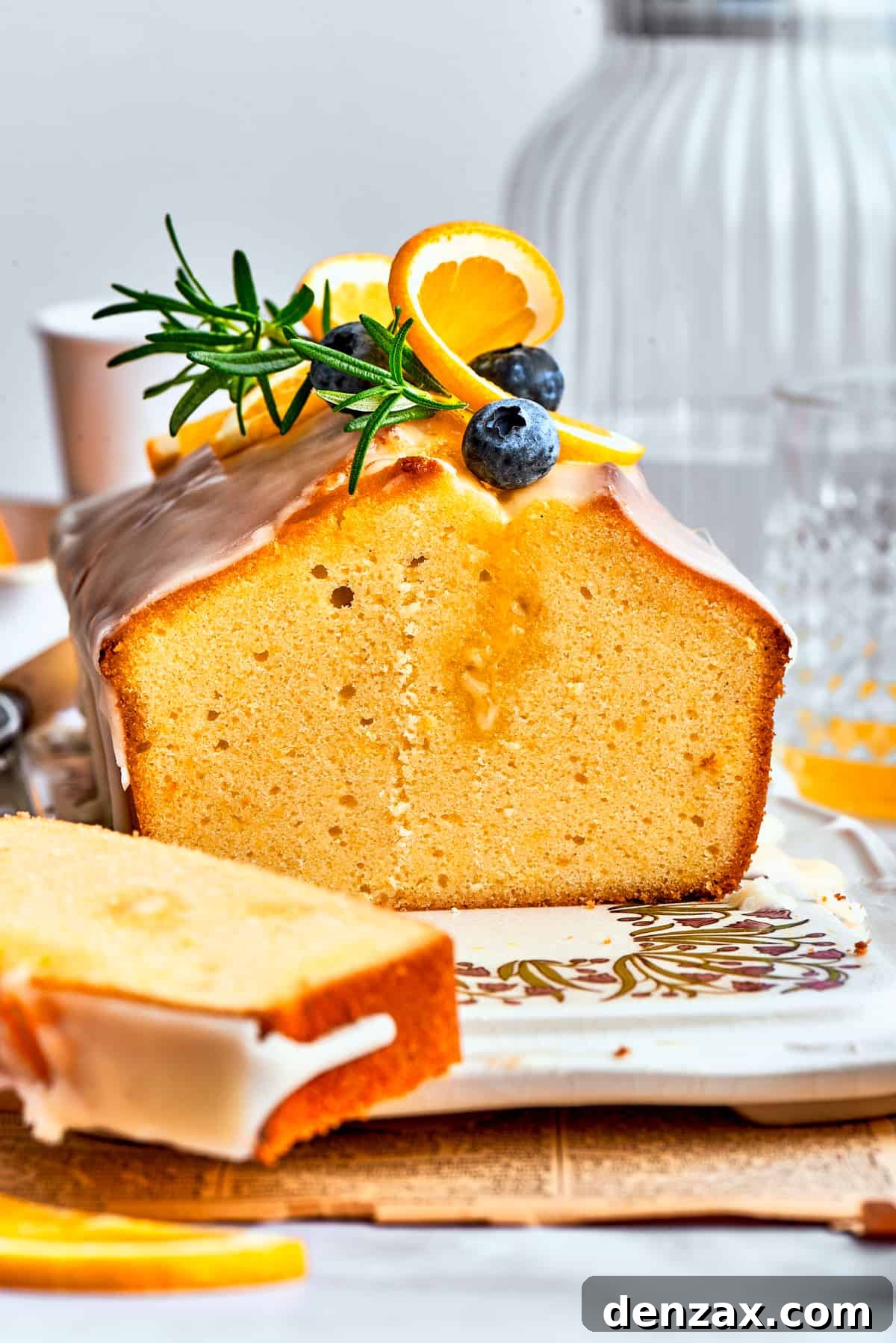 A side view of a slice of orange cake and the entire loaf, topped with blueberries, fresh rosemary, and orange slices.