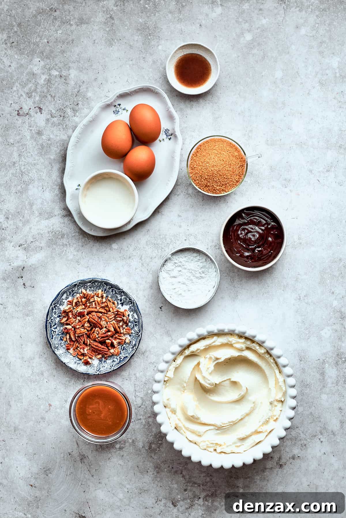 The ingredients for turtle cheesecake are shown portioned out on a white background: eggs. pecans, caramel, cream cheese, chocolate chips, brown sugar, vanilla, sour cream.