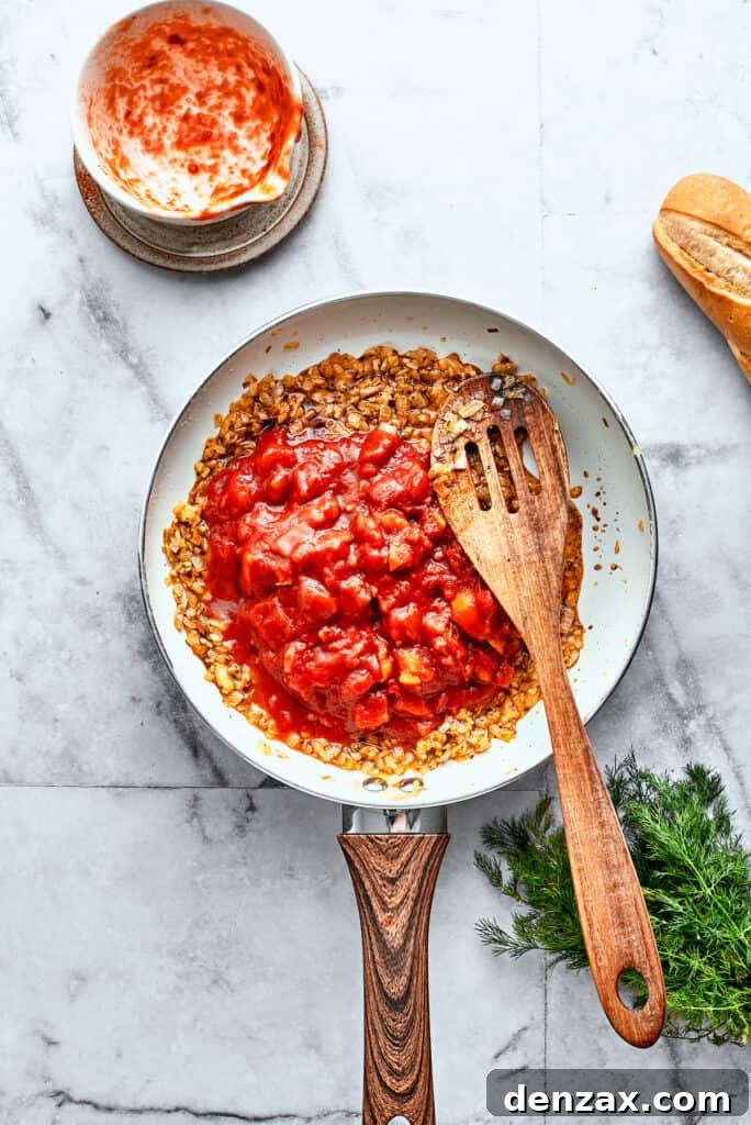 Thick diced tomatoes are stirred into a white skillet with a wooden spoon.