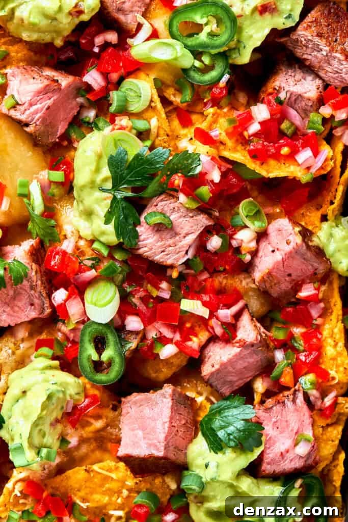 Steak Nachos are piled with homemade guacamole, cheese, pico de Gallo, and juicy steak for a fun twist on nachos. Chunks of juicy steak and peppers top a pan of steak nachos.