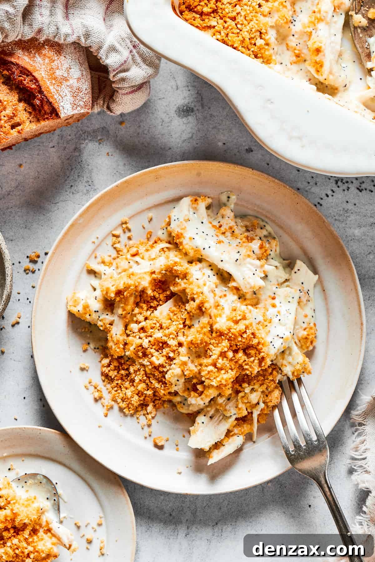 This creamy Poppy Seed Chicken casserole features a rich, poppyseed-flecked sauce that wraps cozily around tender shredded chicken. A casserole dish of poppy seed chicken is shown on a table with a loaf of bread and a plate holding a serving of the chicken.