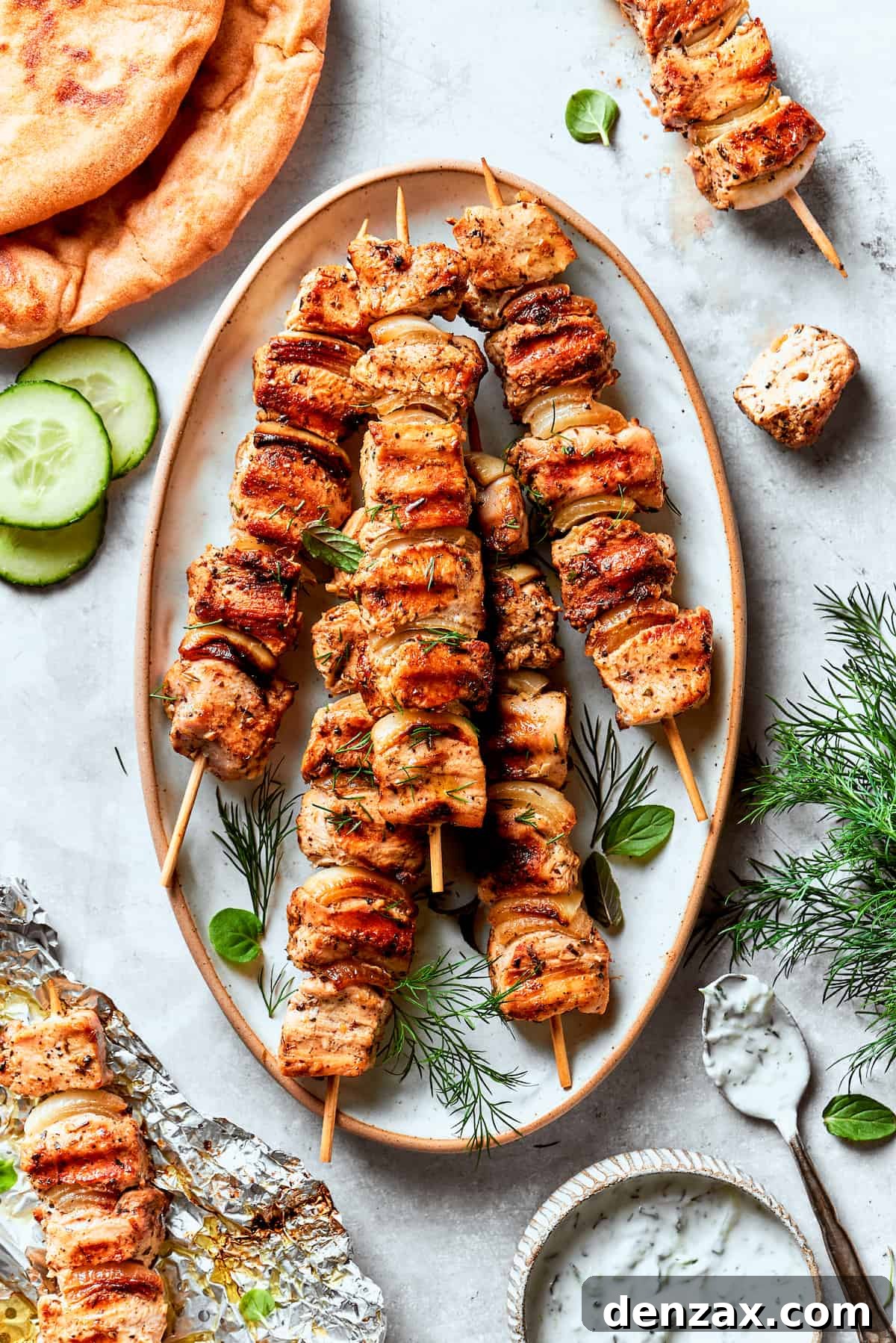 A platter of grilled chicken souvlaki on skewers surrounded by herbs and cucumber slices.