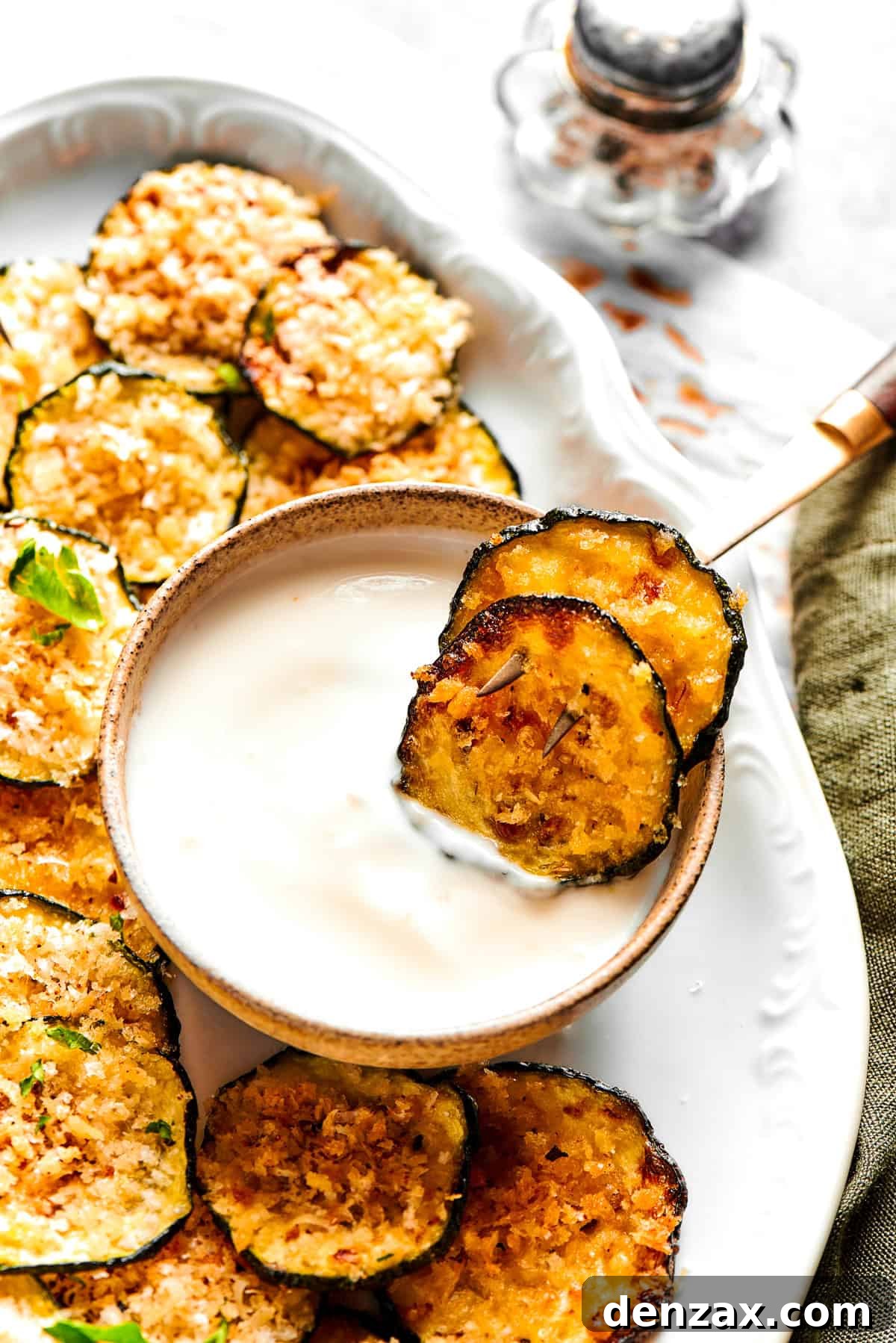 Two crispy zucchini chips in a bowl of yogurt on a platter with more chips.