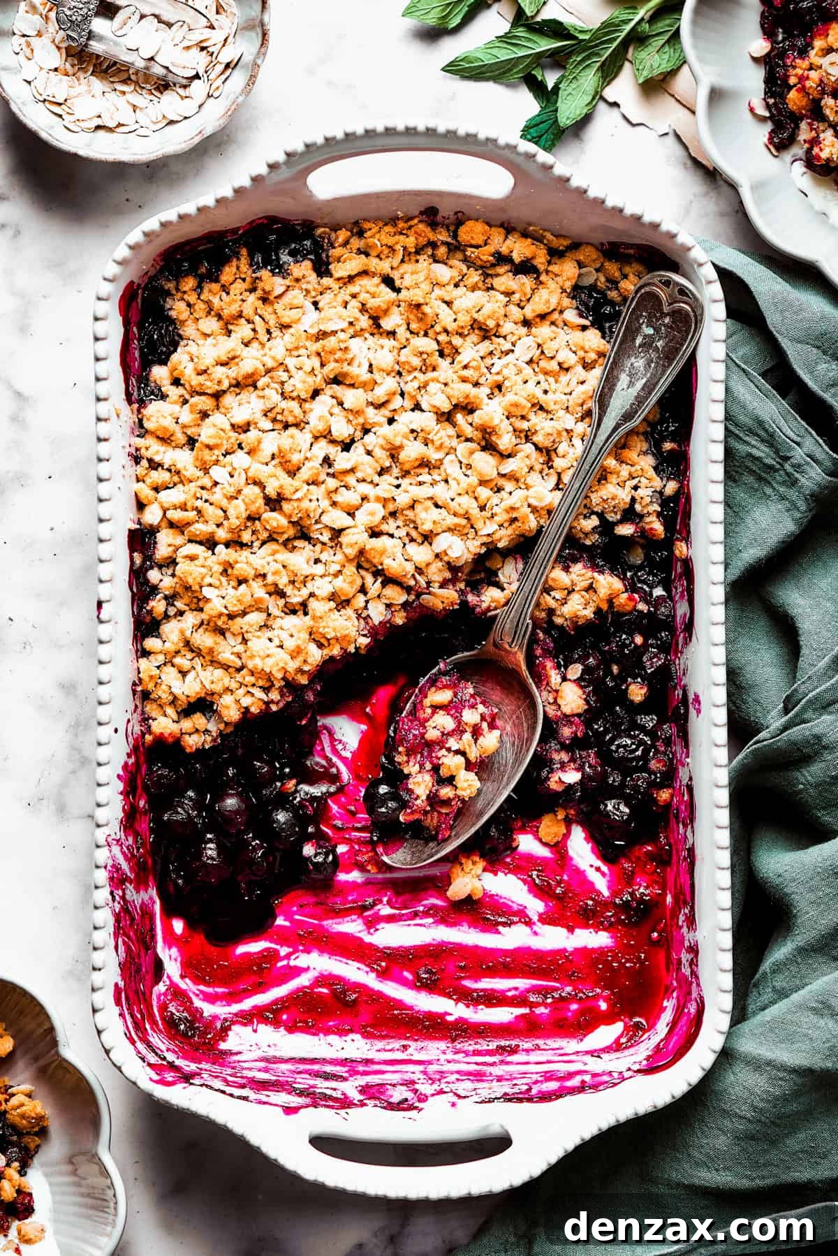Bursting with juicy berries, this Blueberry Crisp Recipe is topped with a buttery brown sugar and oat crumble. Overhead image of a blueberry crisp in a baking dish, with half of the crisp scooped out.