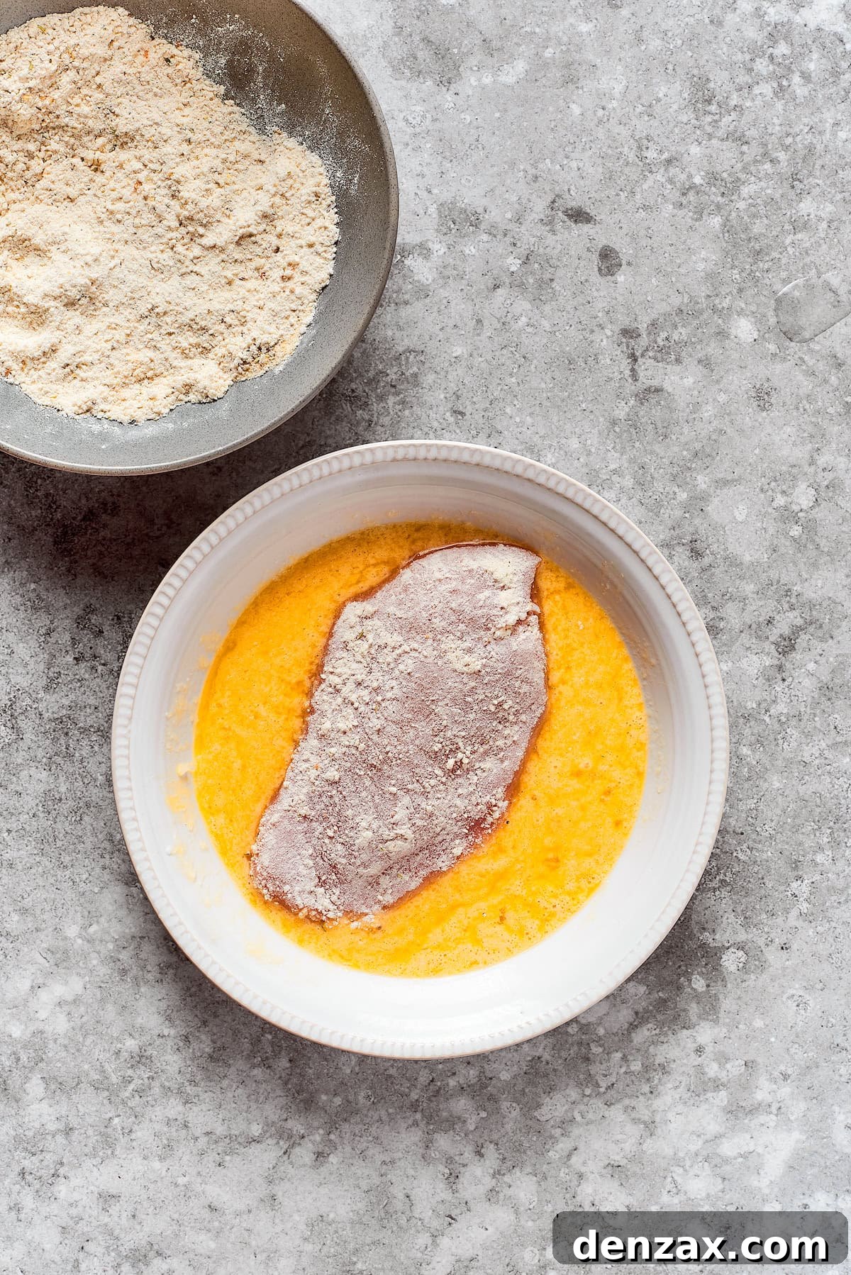 A chicken breast is placed in a bowl of egg wash.