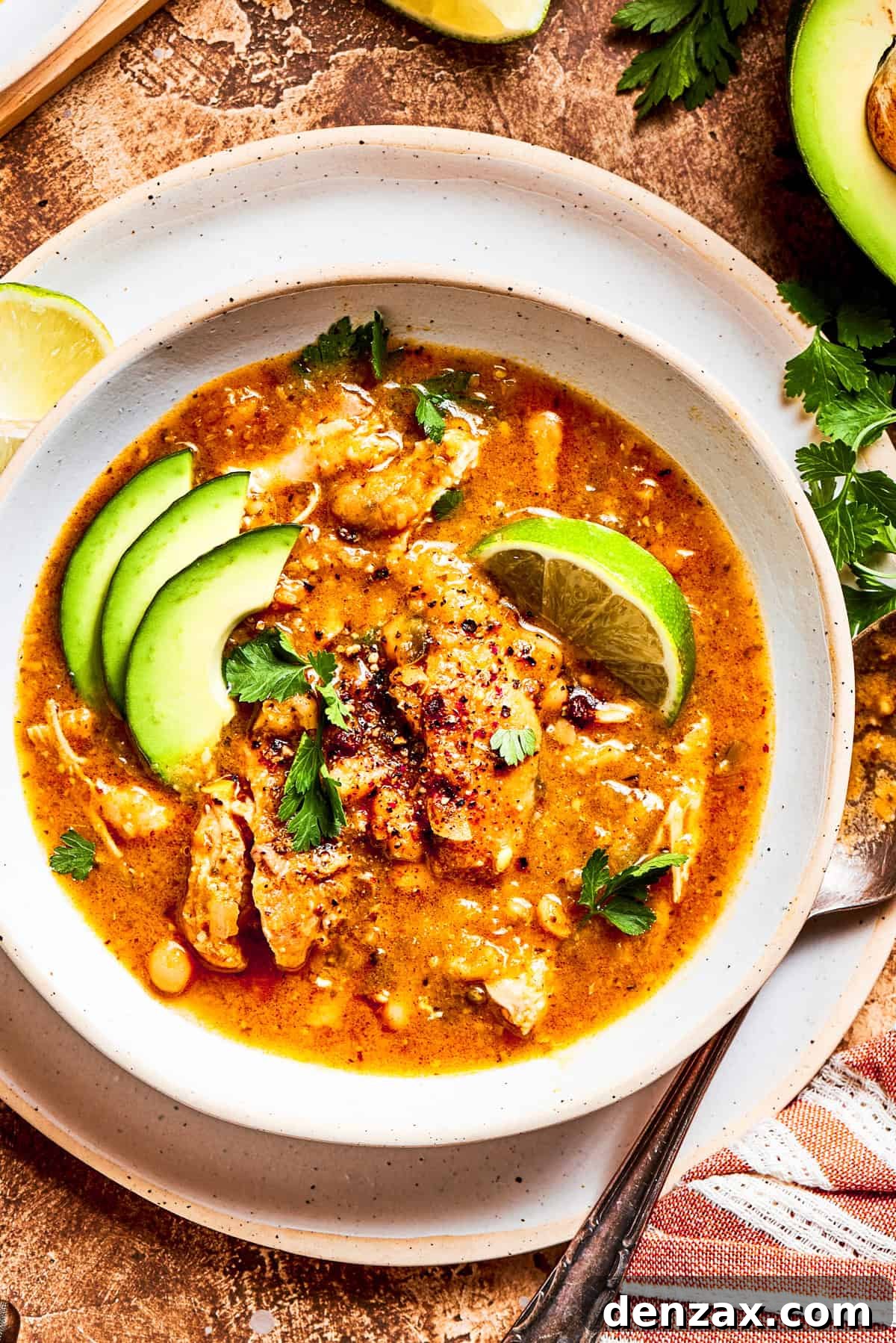 Spicy chicken soup in a bowl garnished with avocado slices and lemon wedges.