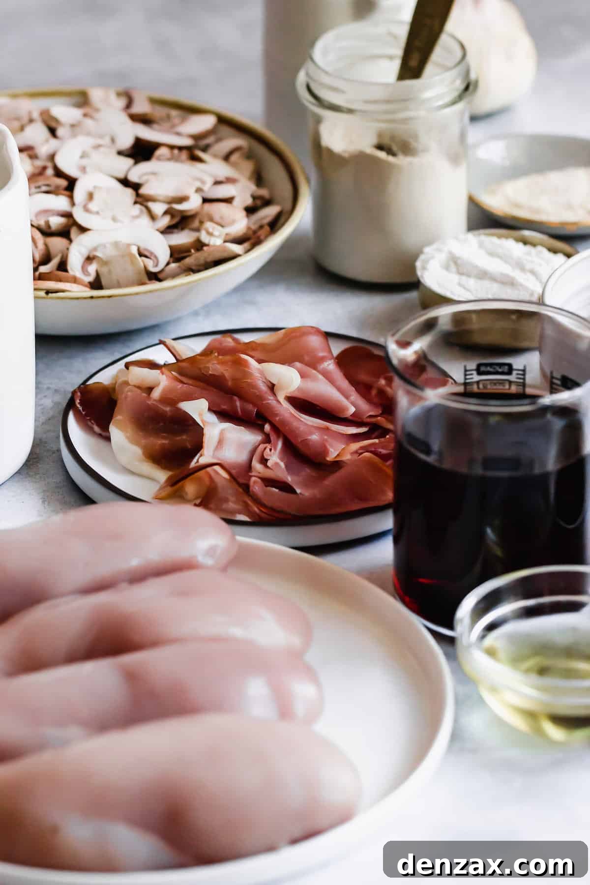 A plate of sliced white mushrooms, a plate of proscuitto, a cup of flour, and a cup of marsala wine.