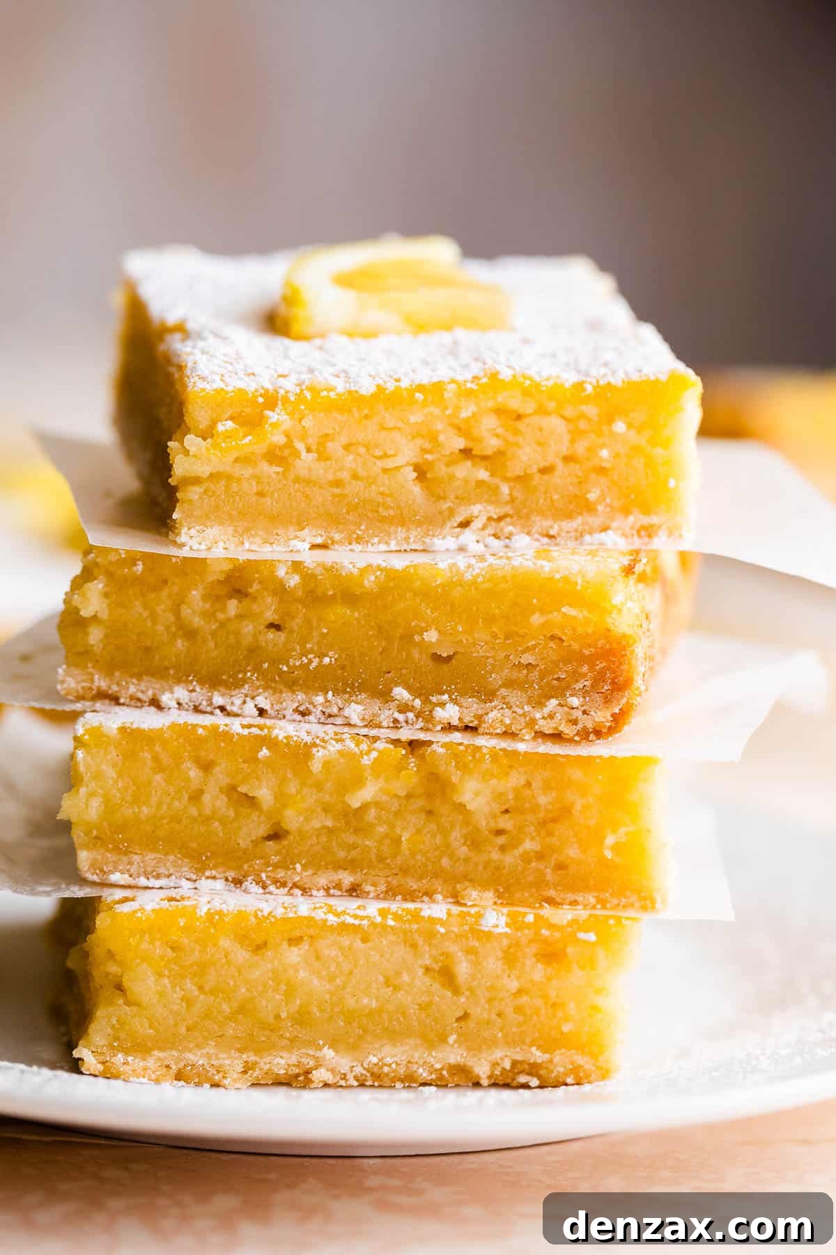 four lemon bars stacked one on top of the other and finished with a dusting of powdered sugar and a slice of lemon on top