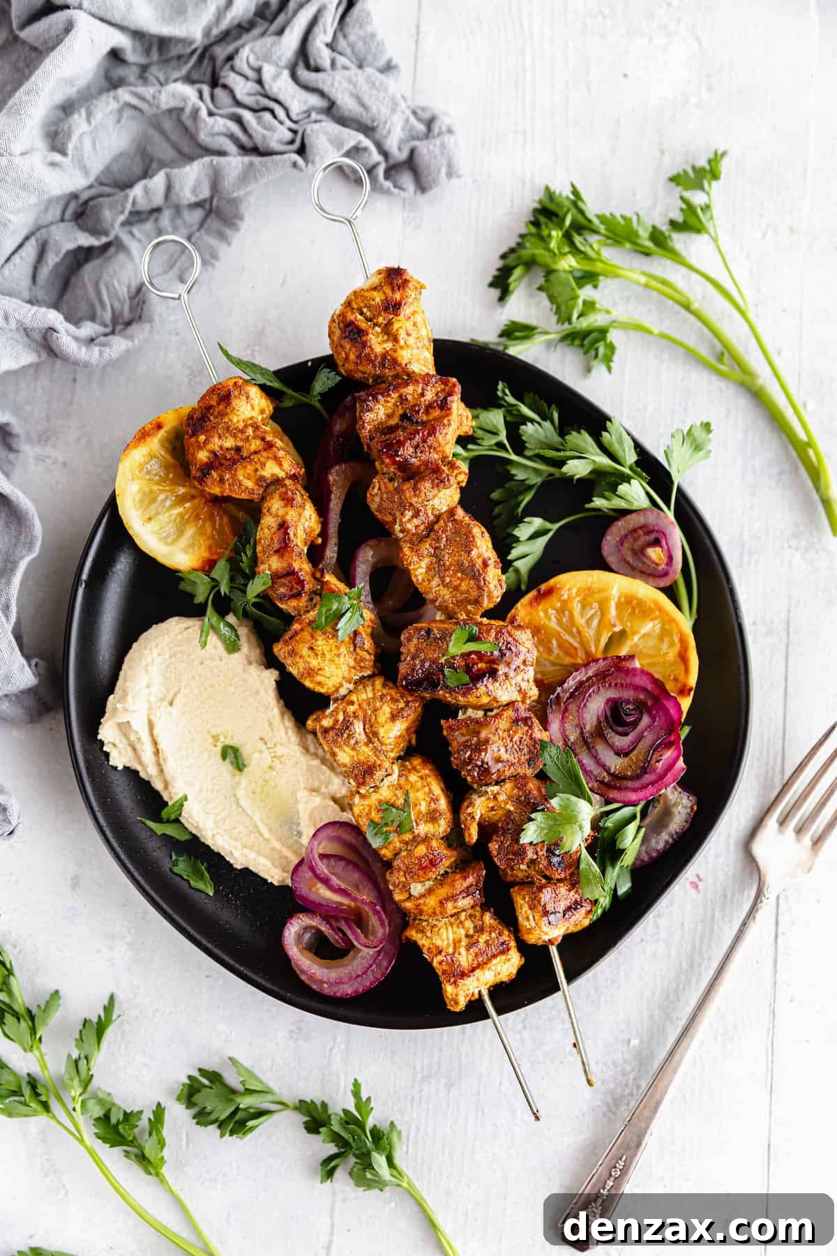 A Black Plate Containing Two Chicken Skewers Along with Various Fruits and Vegetables with a Fork Beside it