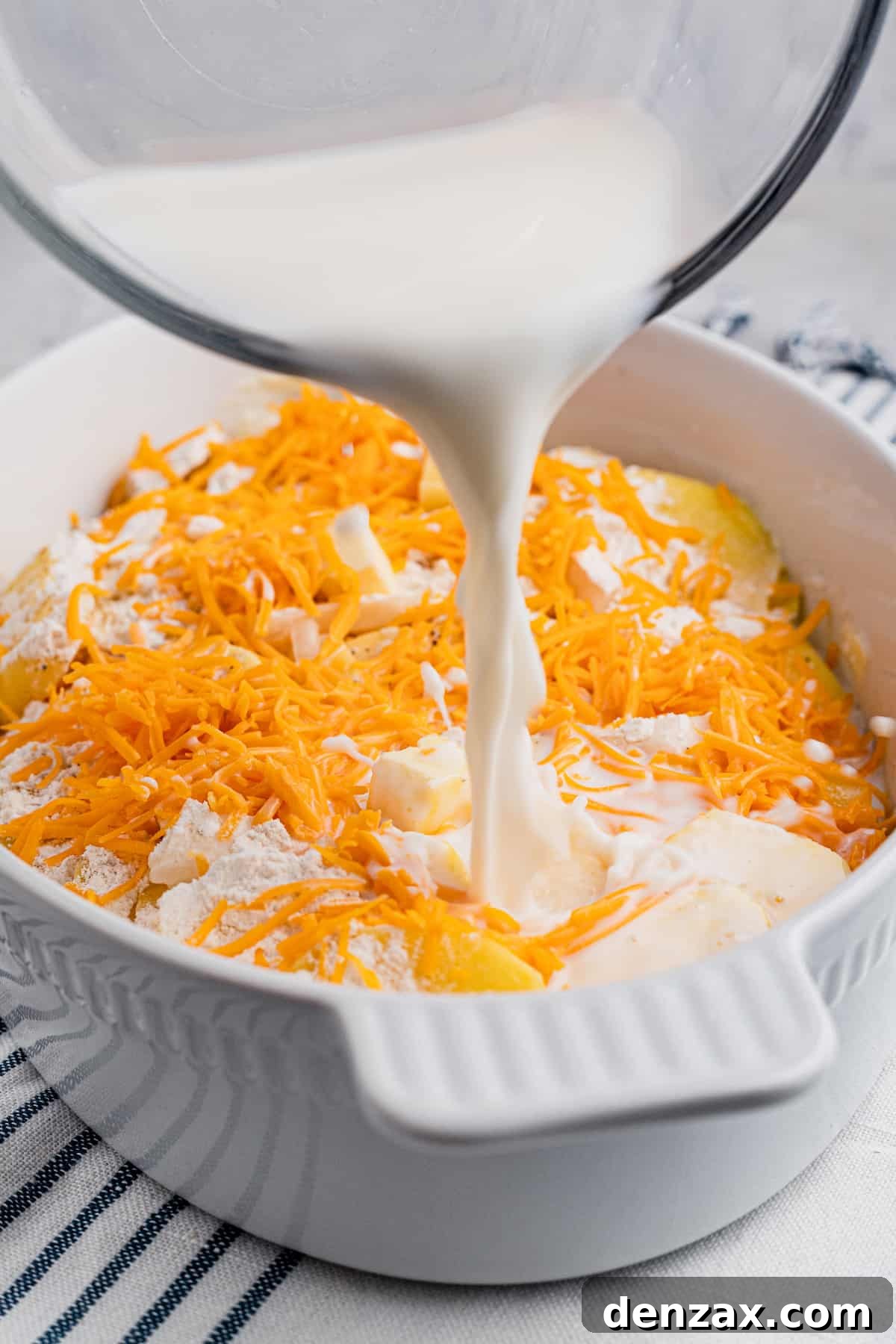 Tender sliced potatoes layered with cheddar cheese and diced onions to make a scalloped potatoes casserole. Pouring milk over sliced potatoes and shredded cheese in a baking dish.