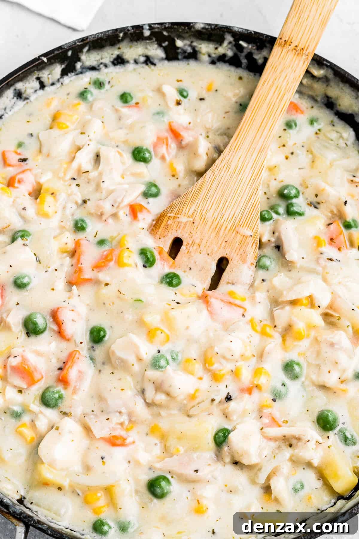 Creamy pot pie filling, featuring carrots, peas, corn, diced potatoes, and chicken, being stirred with a wooden spoon.