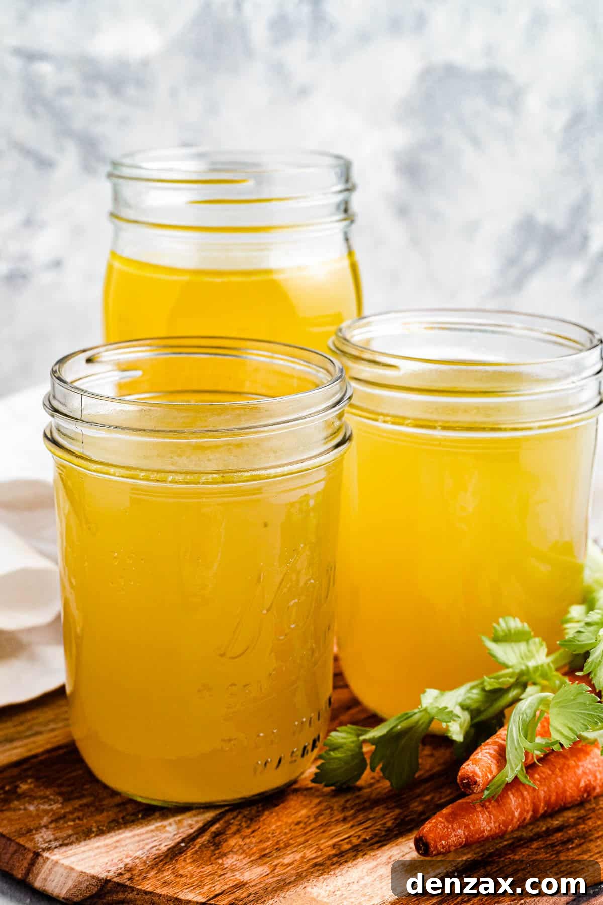 3 jars of homemade chicken broth on a cutting board with raw carrots nearby.