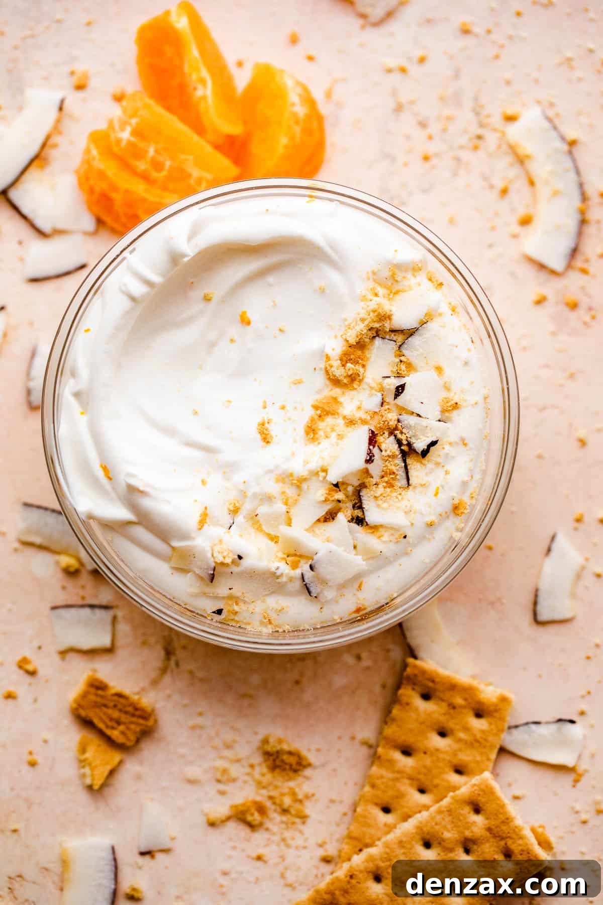 overhead shot of a creamy coconut dip served in a glass bowl with tangerine slices and coconut shavings placed around and on the sides of the bowl.