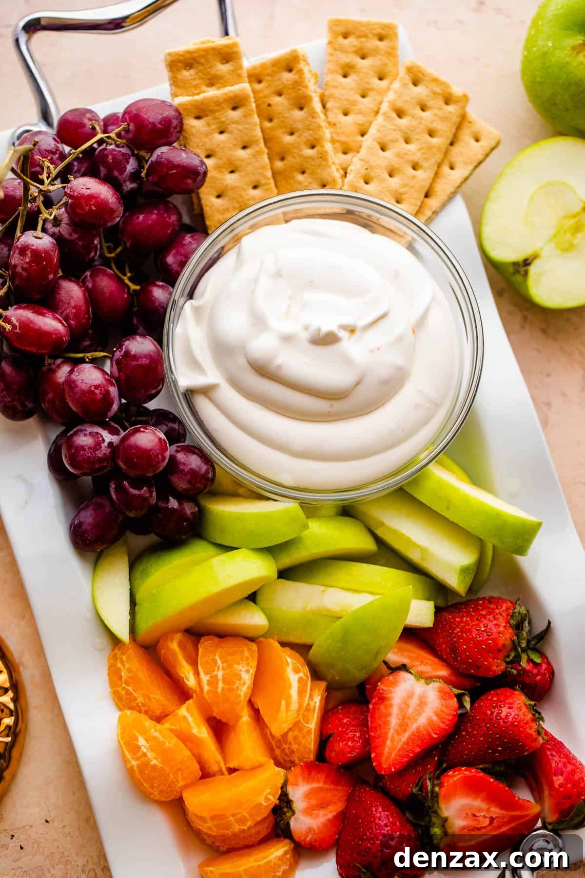 Bowl of creamy coconut fruit dip, green apple slices, tangerine slices, strawberries, grapes, and graham crackers arranged on a long serving plate.