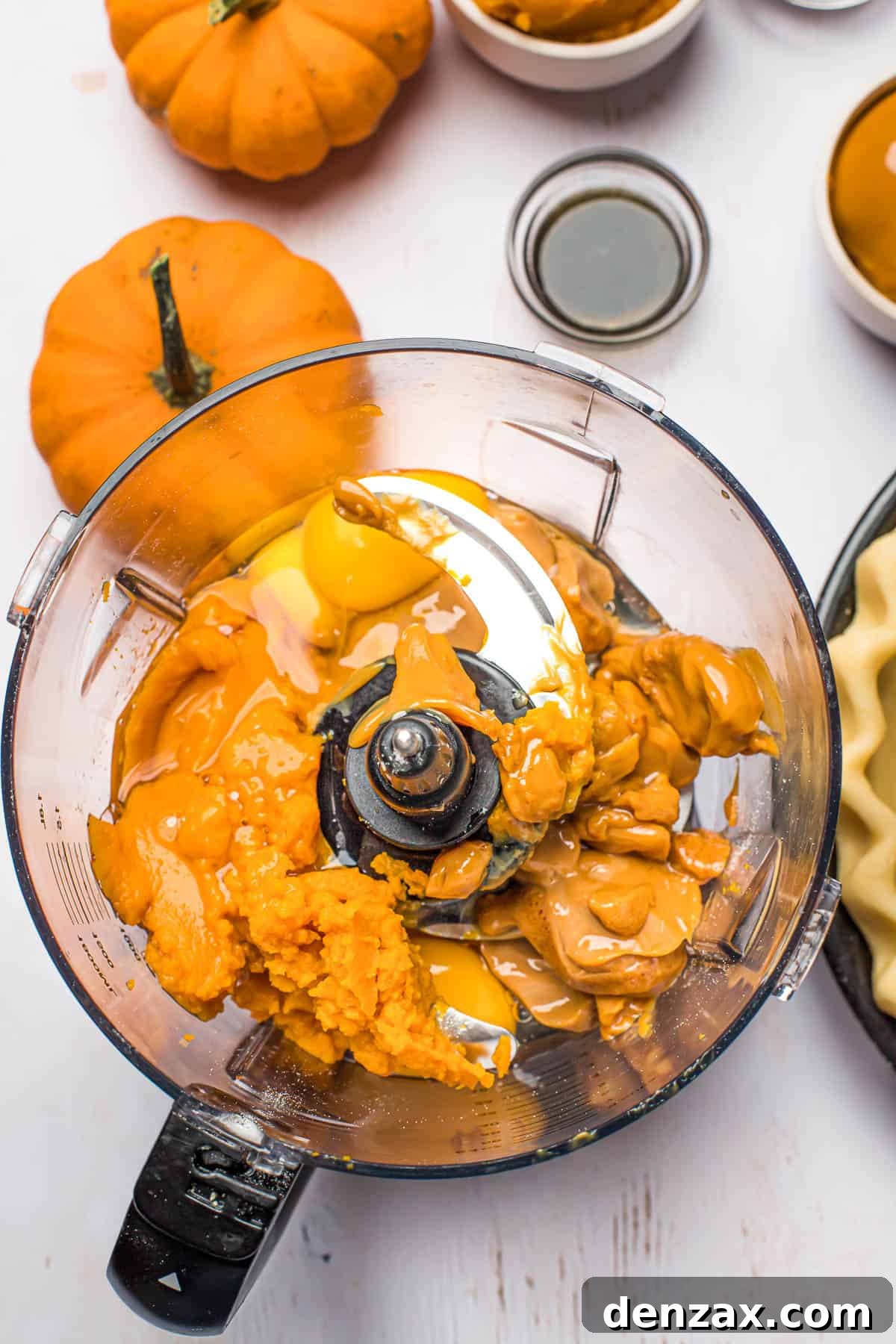 This homemade and easy Pumpkin Pie recipe is a holiday favorite! Everyone will love the tender pie crust, warm spices, and dulce de leche pumpkin filling. Pumpkin filling ingredients in a blender on a white work surface, with mini pumpkins decorating the surface.
