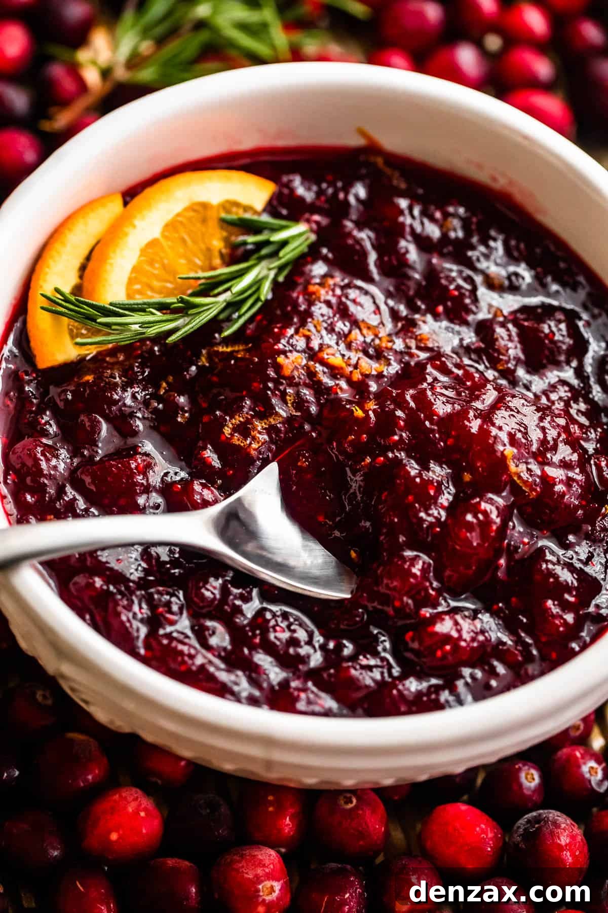 cranberry sauce inside of a white bowl surround by fresh cranberries and garnished with orange slices and rosemary sprig.