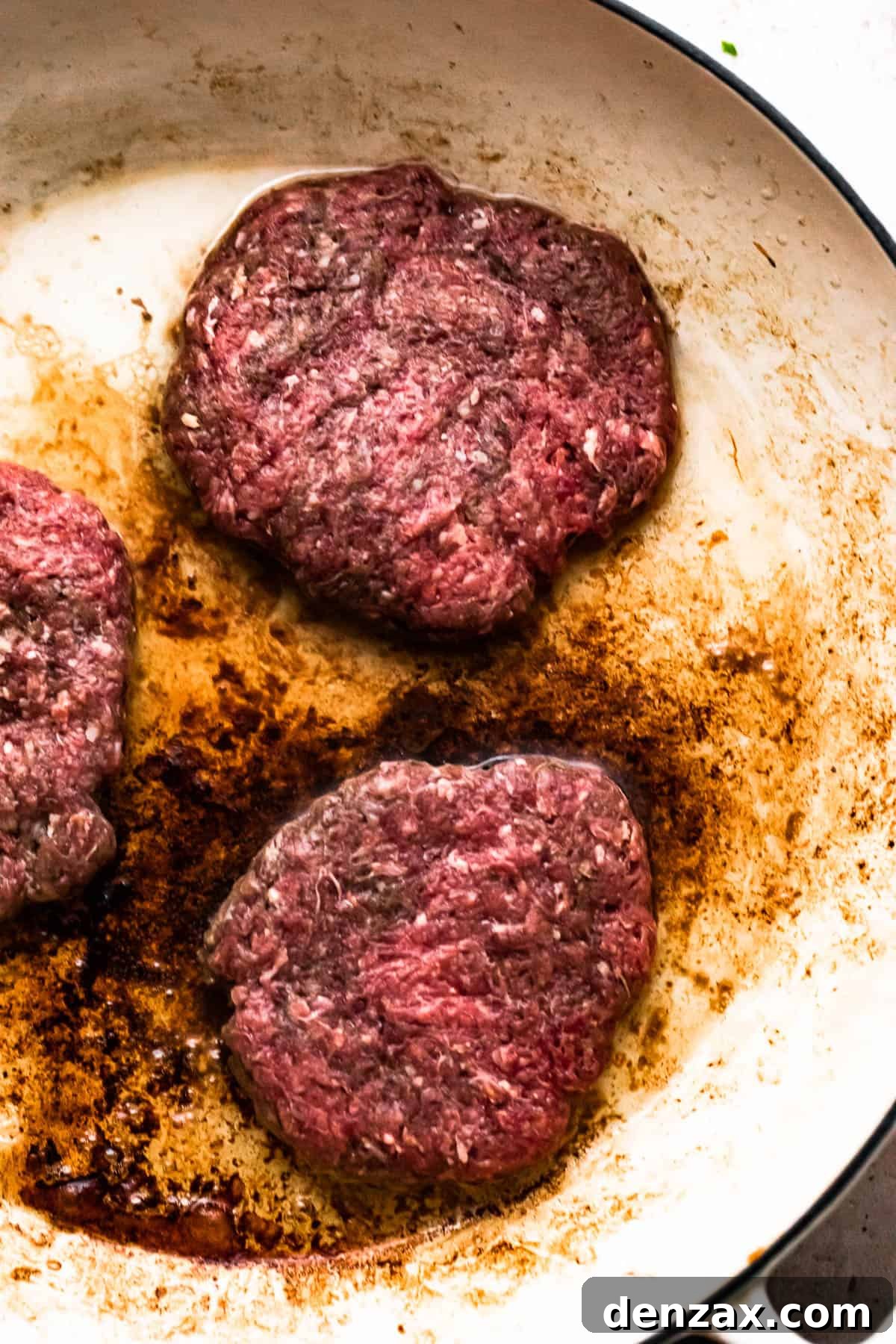 three beef patties cooking in a white skillet.